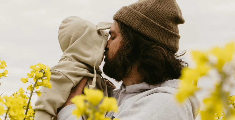 A father wearing a beanie holds up his young child closely to his face, and they're surrounded by yellow flowers