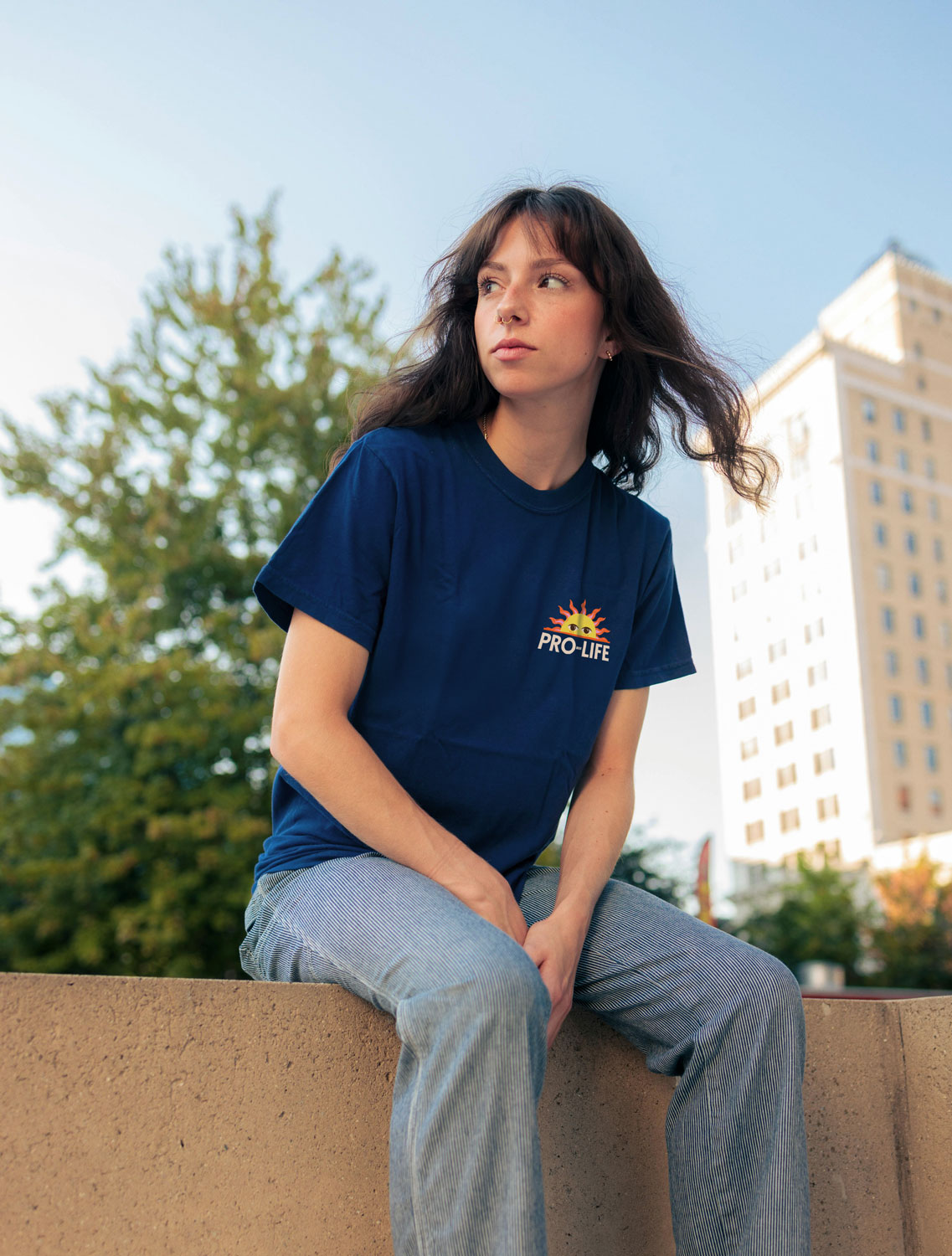 A young woman wearing the navy t-shirt from the ProLife Europe merchandise collection, sitting on a concrete wall in an urban setting